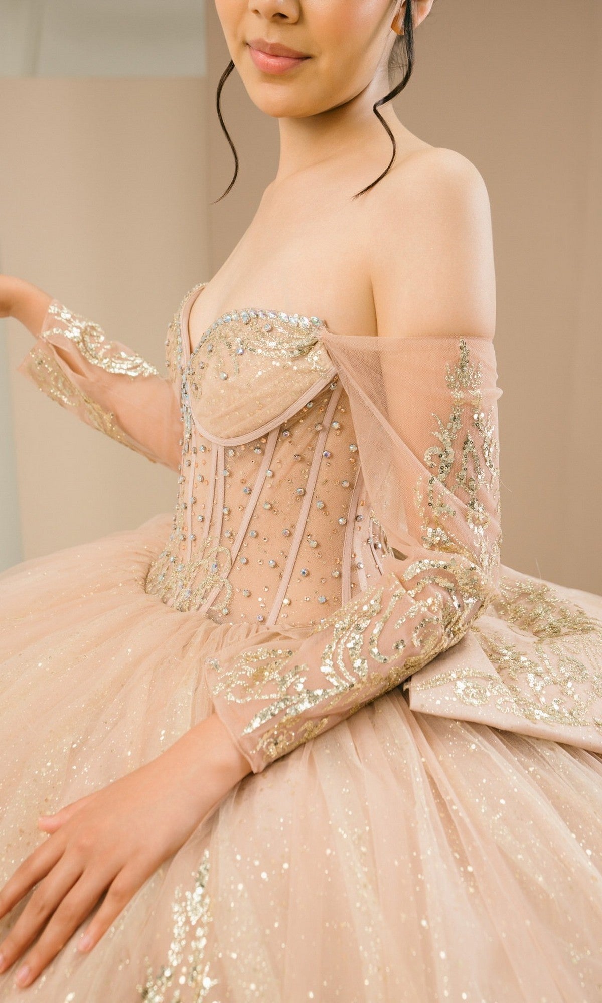 A woman poses elegantly indoors wearing the Quinceanera Dress 8545JQ By Cinderella Couture, an off-the-shoulder ball gown with a sweetheart neckline, gold embroidery, and beaded sequin embellishments.
